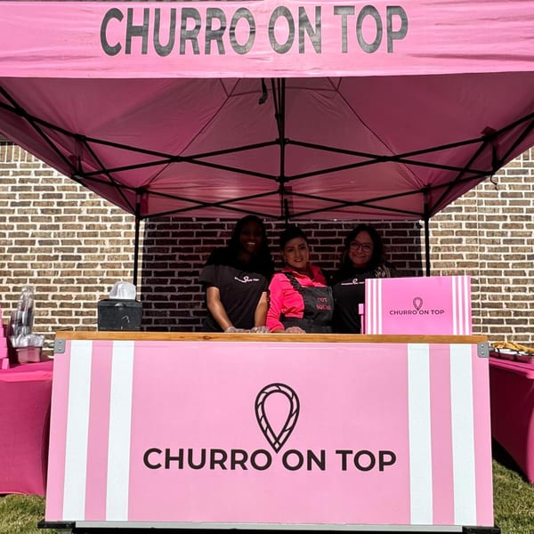 Three women smiling behind a pink "Churro On Top" food stand with a matching tent.