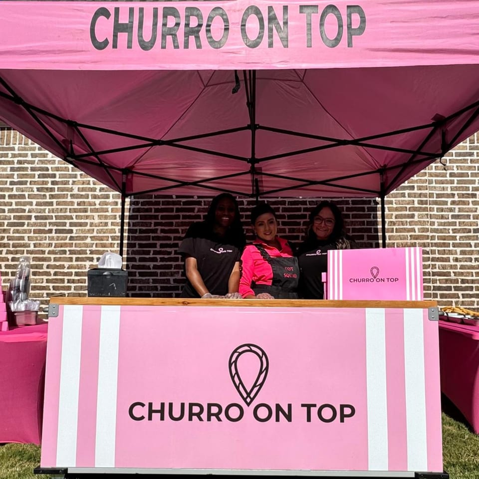 Three women smiling behind a pink "Churro On Top" food stand with a matching tent.