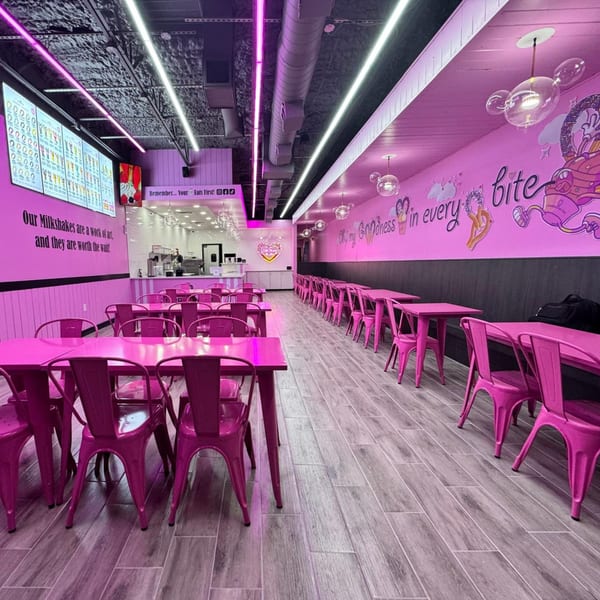 A vibrant pink restaurant interior with rows of tables and chairs, illuminated by neon lights.