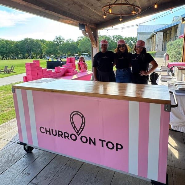 Three women stand behind a pink "Churro On Top" food cart with pink boxes on a patio.