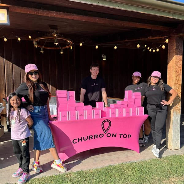 Four adults and one child stand behind a pink "Churro On Top" booth with many pink boxes.